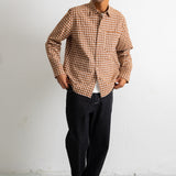 A man wearing a brown and beige checkered long-sleeve button-down shirt, dark pants, and black-and-white sneakers, standing against a plain white wall.