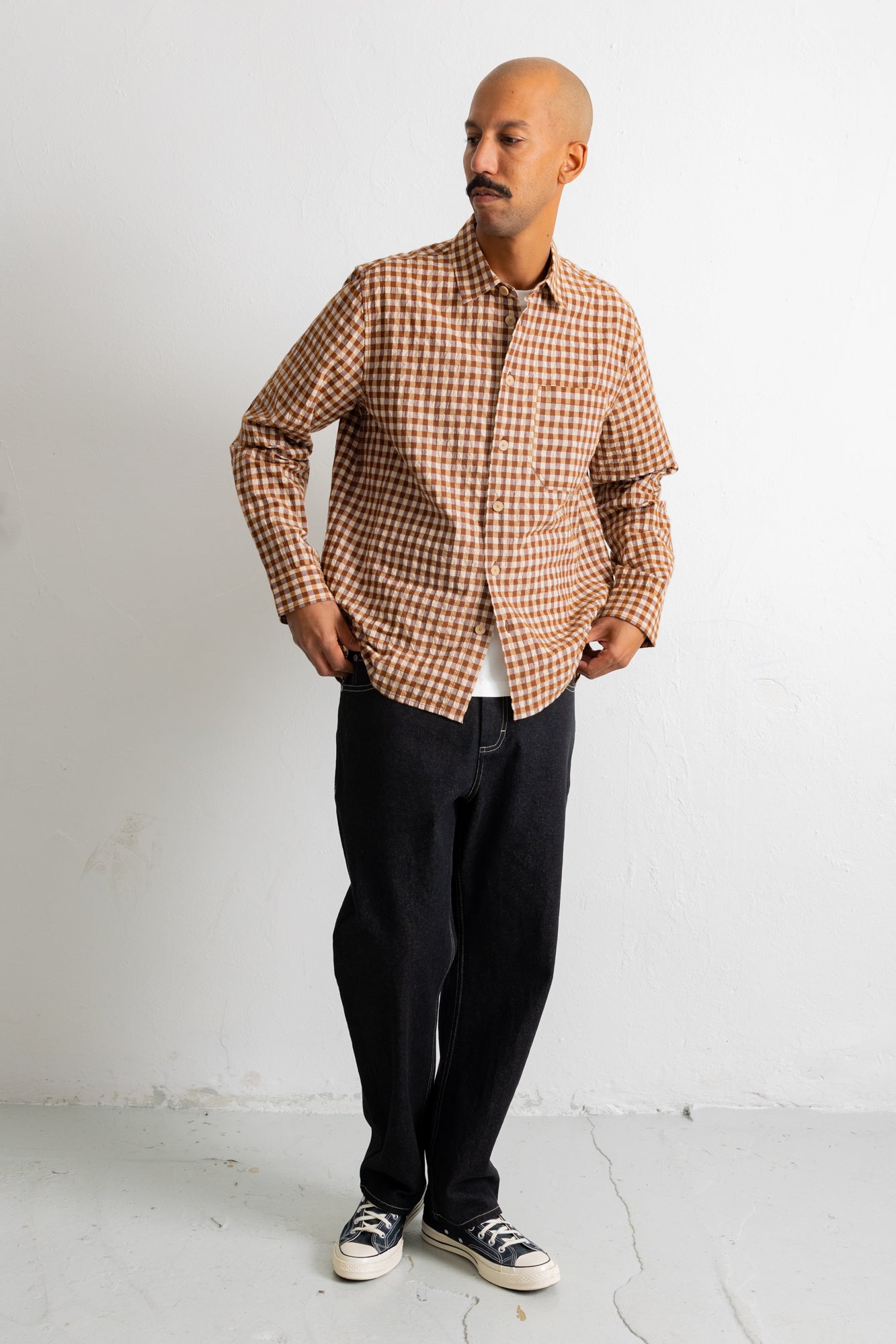 A man wearing a brown and beige checkered long-sleeve button-down shirt, dark pants, and black-and-white sneakers, standing against a plain white wall.