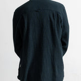 A man viewed from behind wearing a dark navy blue and black checkered long-sleeve shirt and teal jeans, standing against a plain white wall.