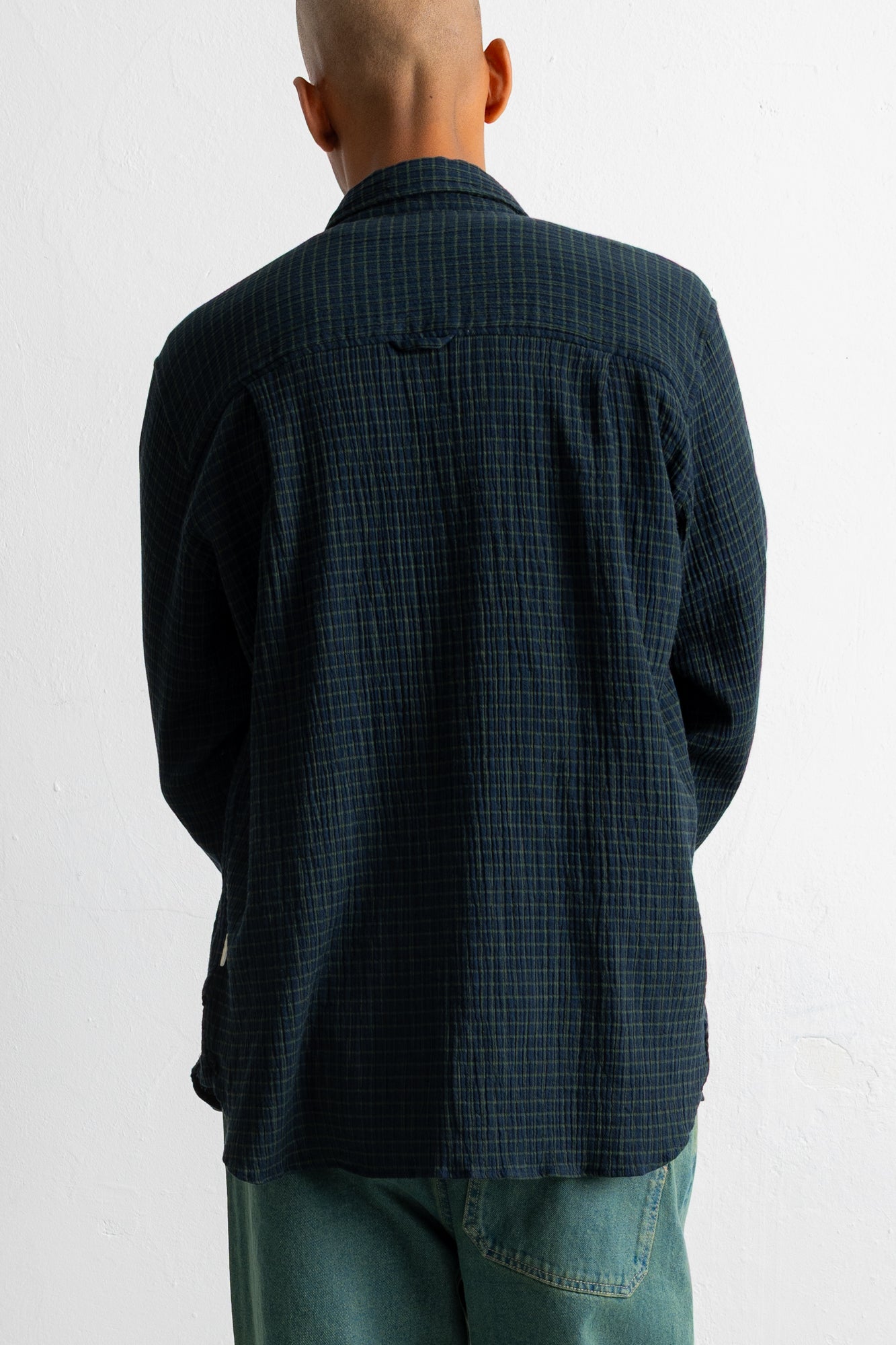 A man viewed from behind wearing a dark navy blue and black checkered long-sleeve shirt and teal jeans, standing against a plain white wall.