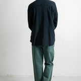 A man viewed from behind wearing a dark navy blue textured long-sleeve button-up shirt and green trousers, standing against a plain white wall.
