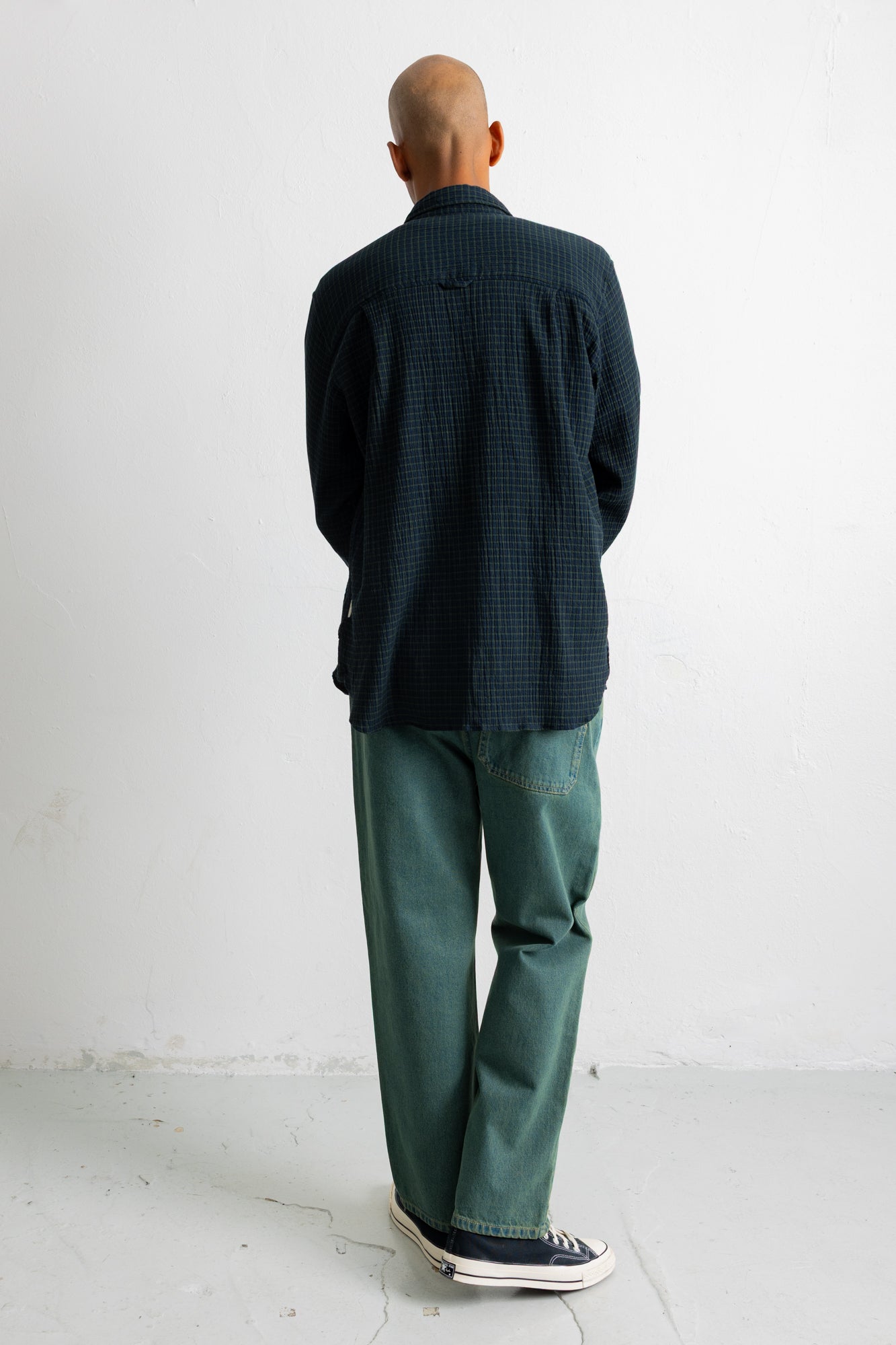 A man viewed from behind wearing a dark navy blue textured long-sleeve button-up shirt and green trousers, standing against a plain white wall.