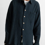 A man wearing a dark navy blue textured button-down shirt with a subtle grid pattern, paired with green pants and a white undershirt, shown from the chin down against a plain white wall.