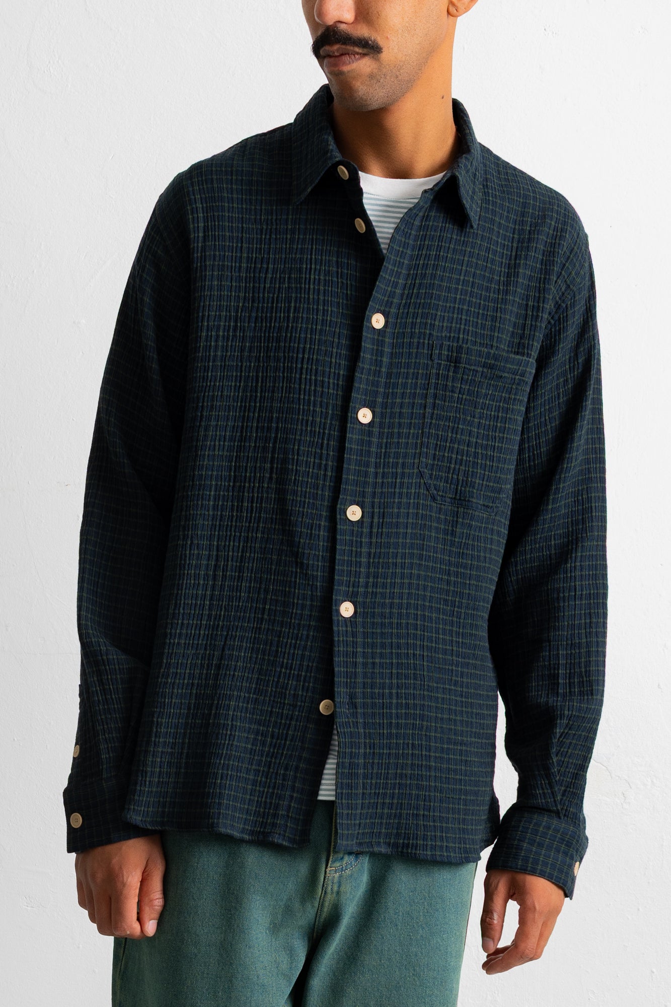 A man wearing a dark navy blue textured button-down shirt with a subtle grid pattern, paired with green pants and a white undershirt, shown from the chin down against a plain white wall.