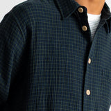 Close-up of a man wearing a dark navy plaid button-down shirt over a white undershirt, showing the collar, buttons, and textured fabric.