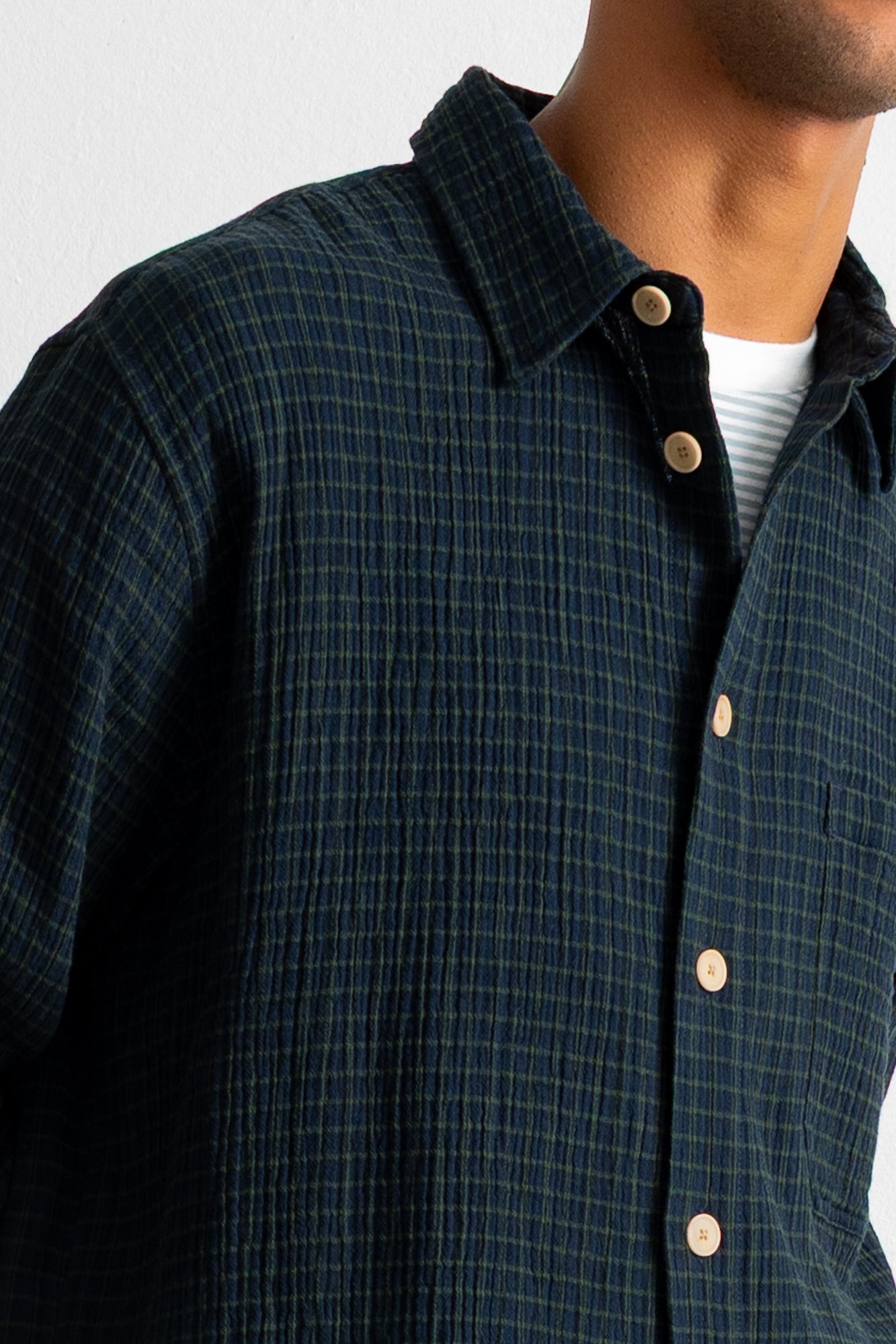 Close-up of a man wearing a dark navy plaid button-down shirt over a white undershirt, showing the collar, buttons, and textured fabric.