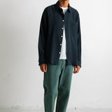A man stands against a white wall wearing a dark navy textured button-up shirt over a white t-shirt, paired with green trousers and black-and-white sneakers.
