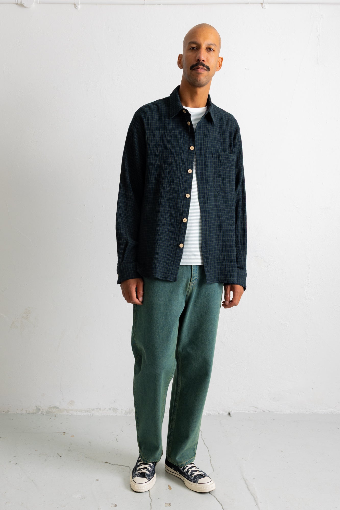 A man stands against a white wall wearing a dark navy textured button-up shirt over a white t-shirt, paired with green trousers and black-and-white sneakers.