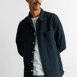 A man wearing a dark navy blue textured overshirt over a white striped t-shirt and teal jeans, standing against a plain white wall.