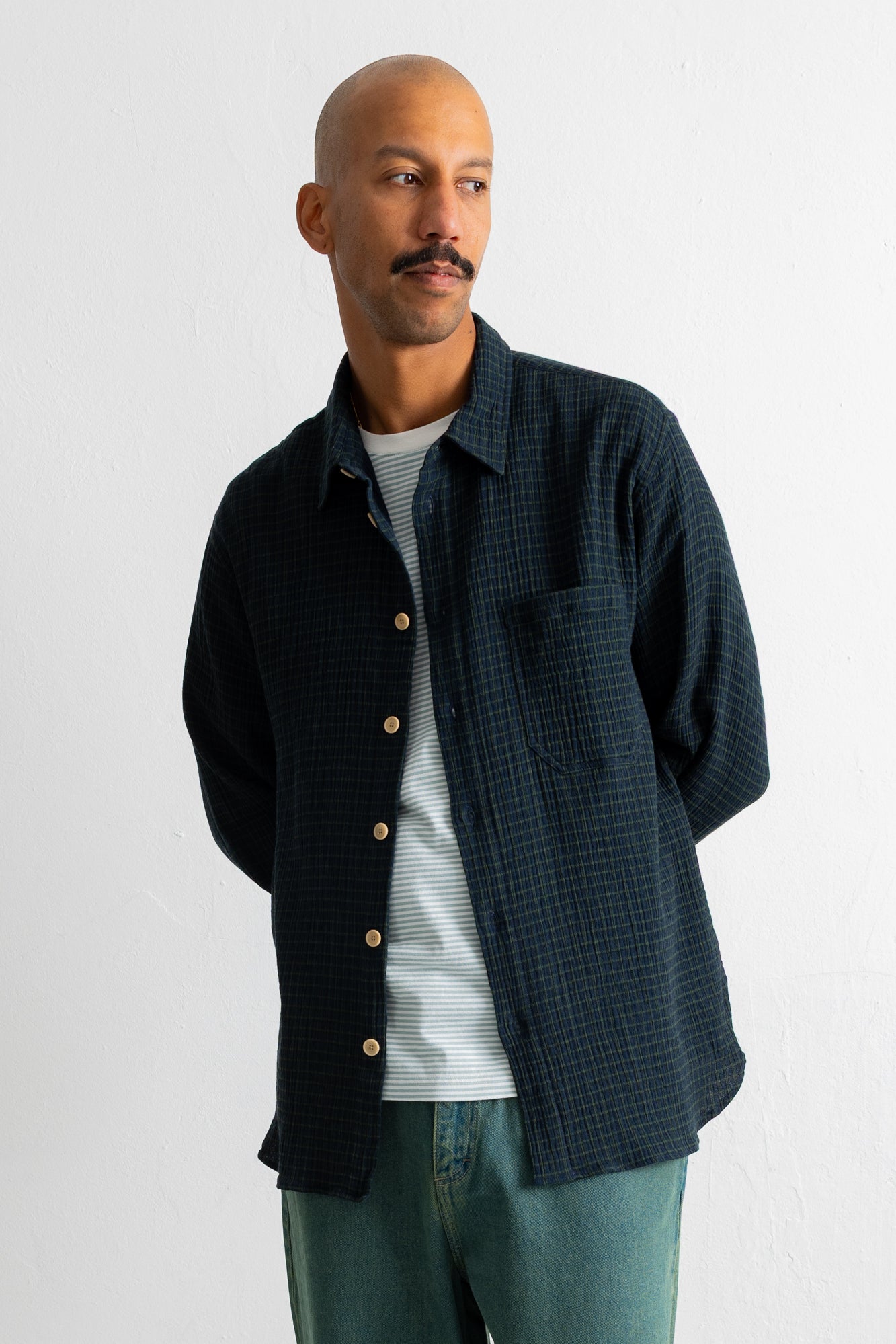 A man wearing a dark navy blue textured overshirt over a white striped t-shirt and teal jeans, standing against a plain white wall.
