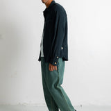 A man wearing a dark navy textured button-up shirt, green wide-leg trousers, and black canvas sneakers, standing in profile against a white studio wall.