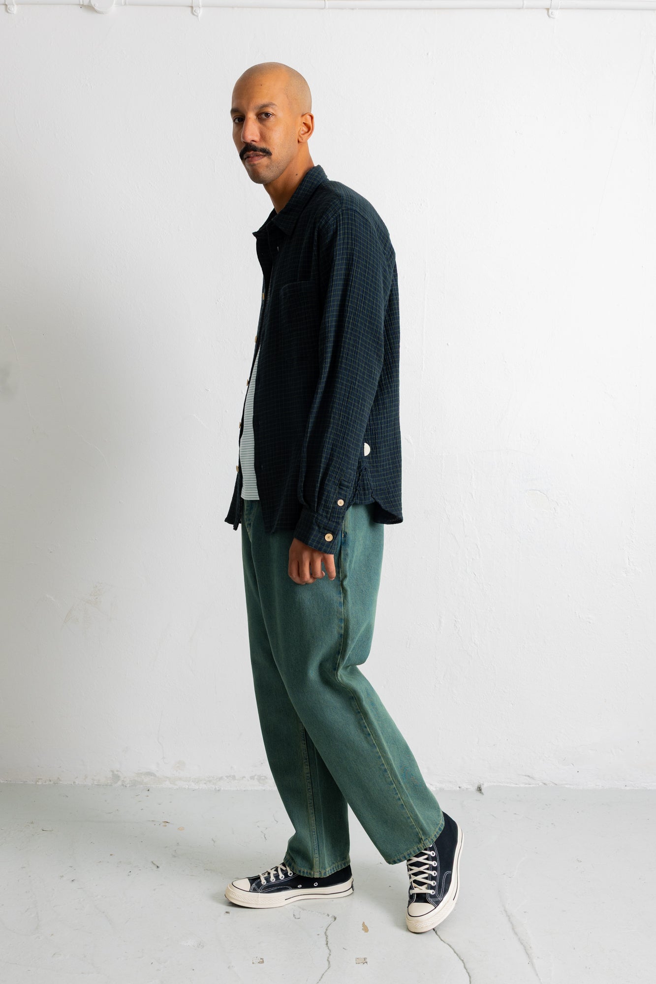 A man wearing a dark navy textured button-up shirt, green wide-leg trousers, and black canvas sneakers, standing in profile against a white studio wall.