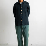 A man stands against a plain white wall wearing a dark navy textured button-up shirt, green trousers, and black canvas sneakers.
