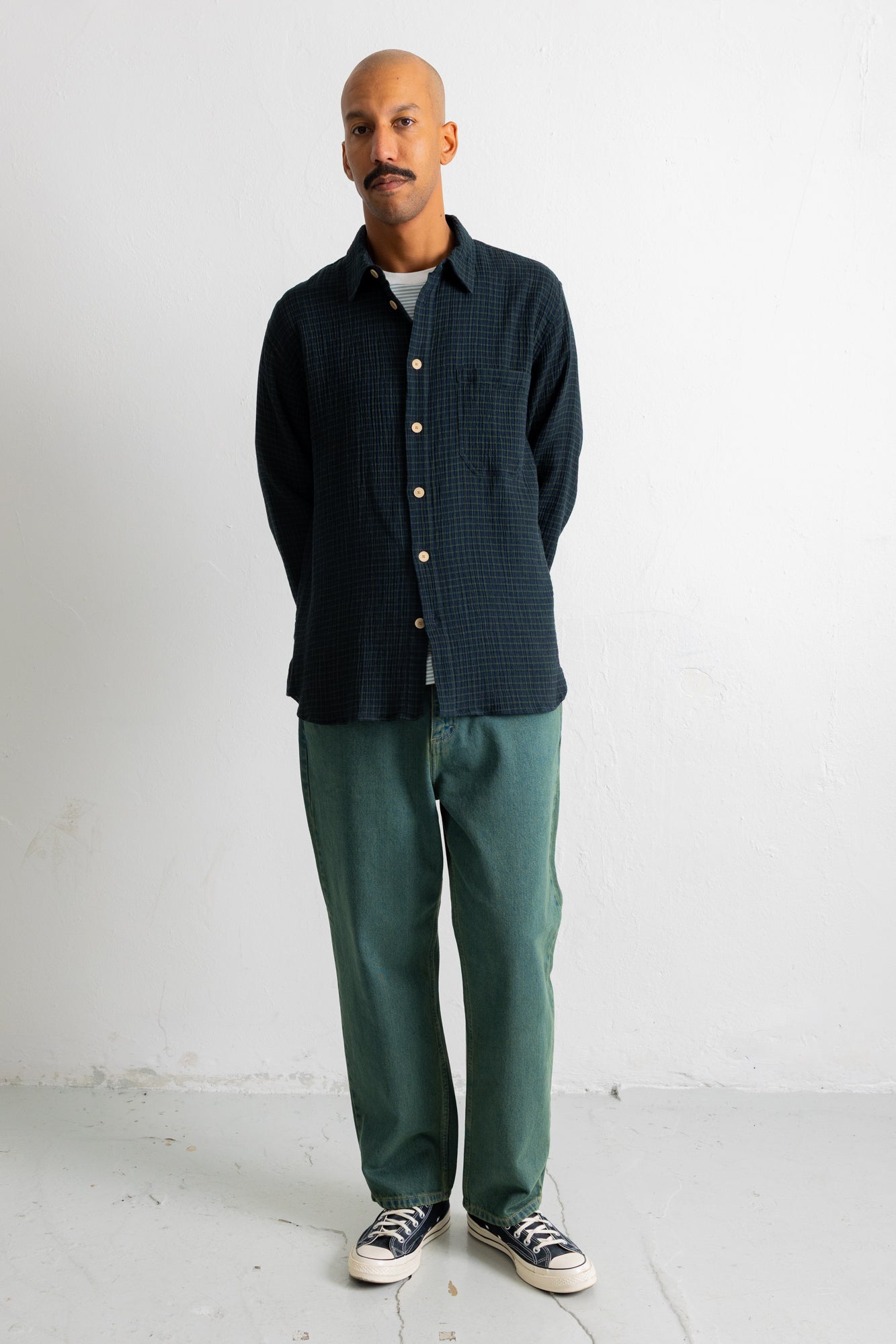 A man stands against a plain white wall wearing a dark navy textured button-up shirt, green trousers, and black canvas sneakers.