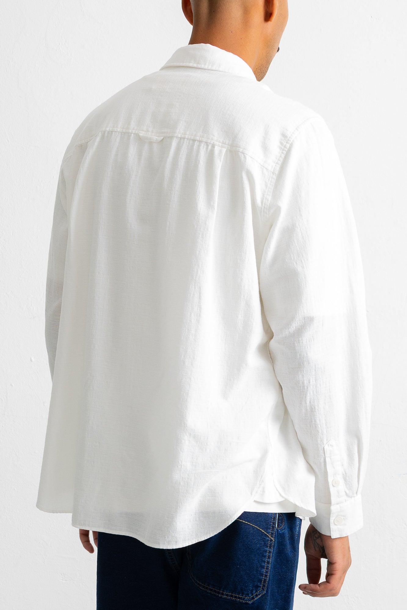 A man viewed from the back wearing a white long-sleeve button-up shirt and dark blue jeans against a plain white background.