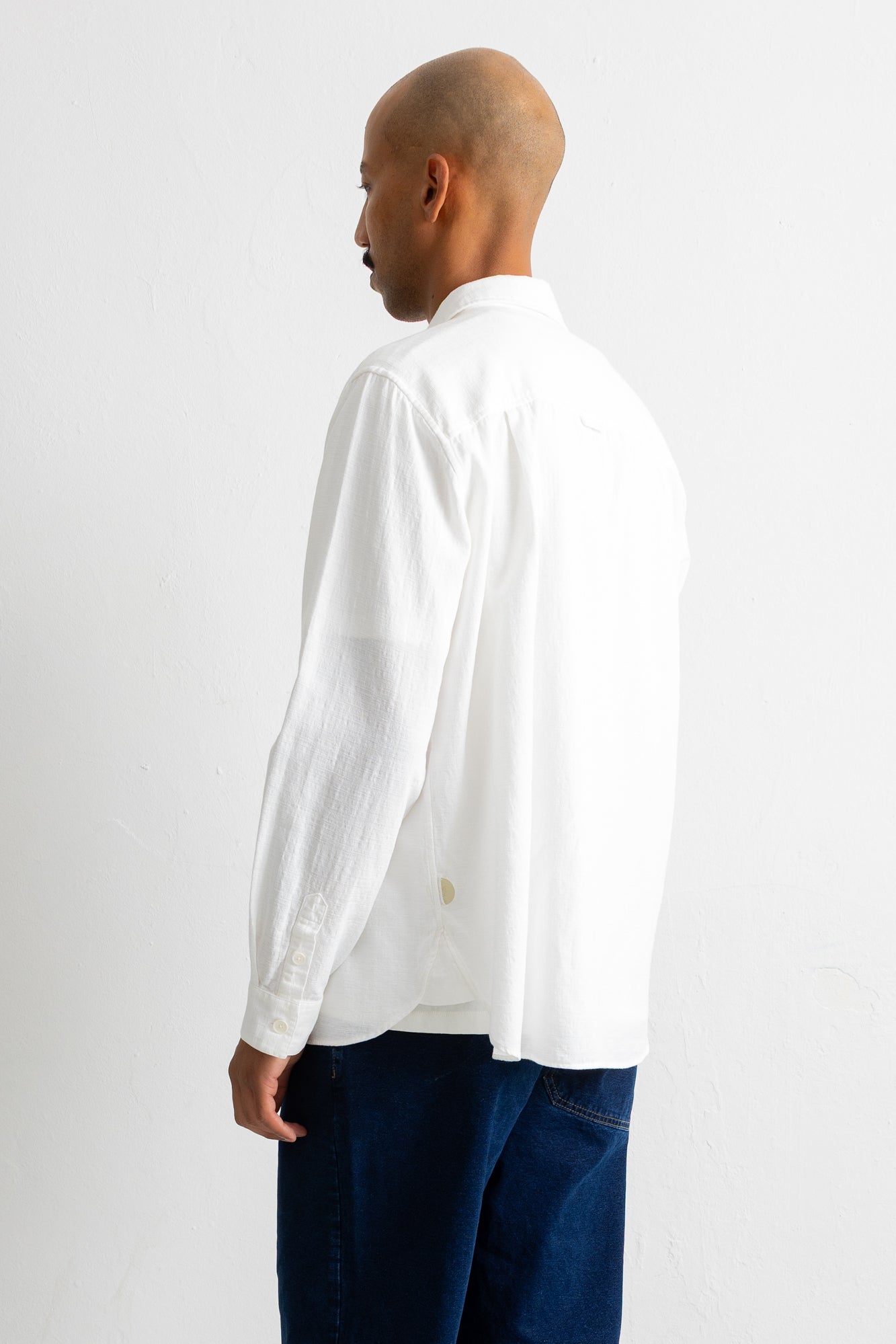A man shown from the back wearing a white long-sleeve linen shirt and dark blue jeans, standing against a plain white studio background.