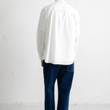 A man viewed from behind wearing a white long-sleeve button-up shirt, dark blue jeans, and black canvas sneakers, standing against a plain white wall.