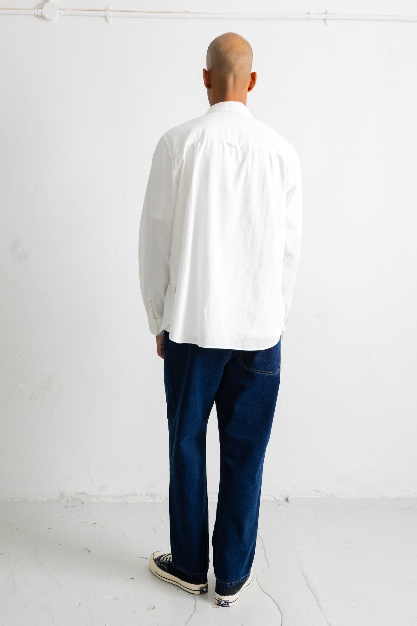 A man viewed from behind wearing a white long-sleeve button-up shirt, dark blue jeans, and black canvas sneakers, standing against a plain white wall.
