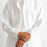 A man wearing a white long-sleeve button-down shirt over a white t-shirt and dark blue jeans, shown from the chin down against a plain white background.
