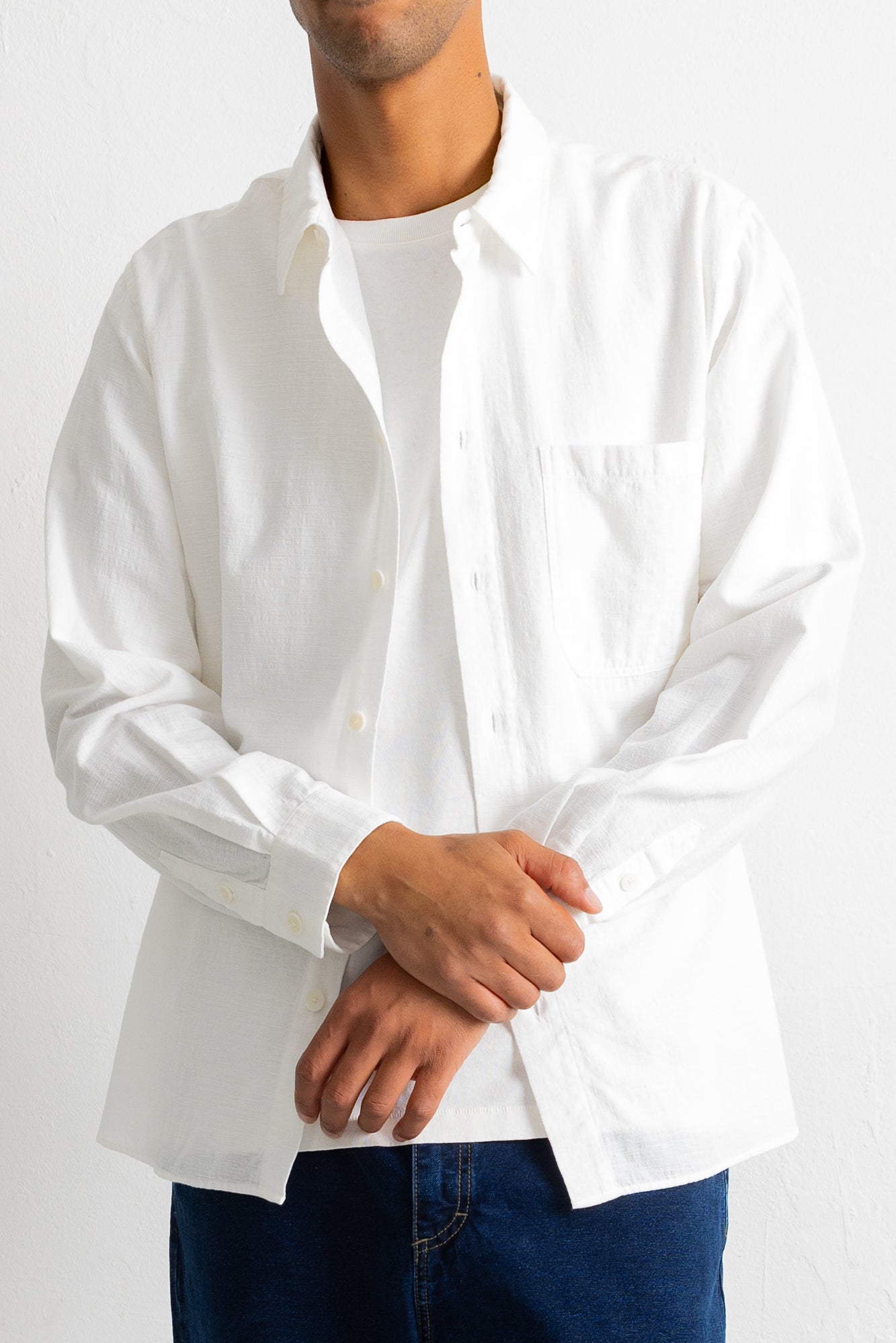 A man wearing a white long-sleeve button-down shirt over a white t-shirt and dark blue jeans, shown from the chin down against a plain white background.