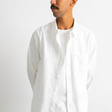 A man wearing a white long-sleeve linen button-up shirt over a white t-shirt and dark blue jeans, standing against a plain white studio background.