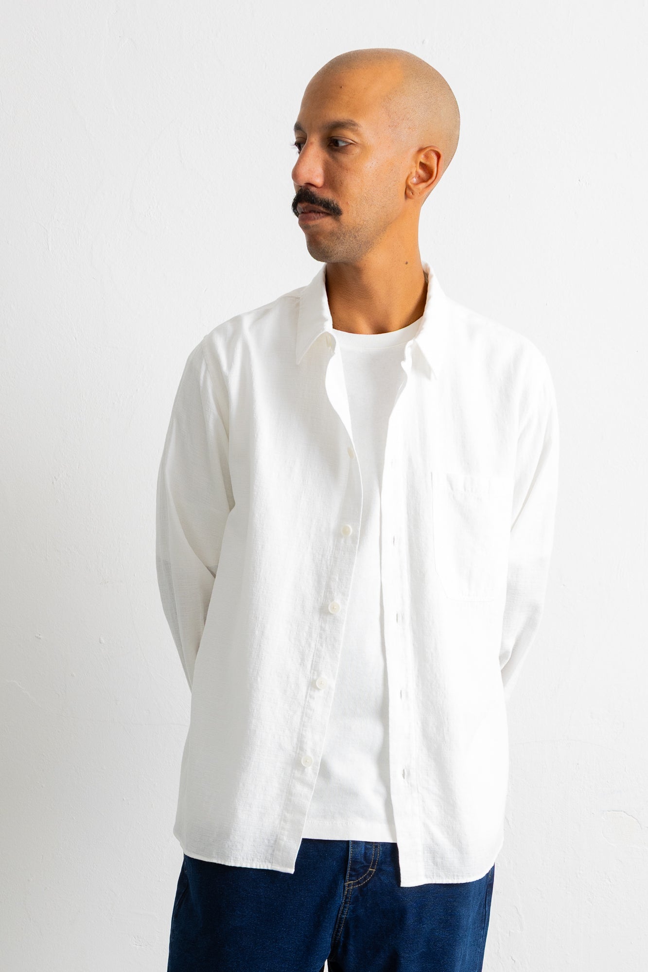 A man wearing a white long-sleeve linen button-up shirt over a white t-shirt and dark blue jeans, standing against a plain white studio background.