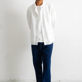 A man wearing a white long-sleeve button-up shirt, dark navy pants, and black-and-white sneakers, standing against a plain white wall.