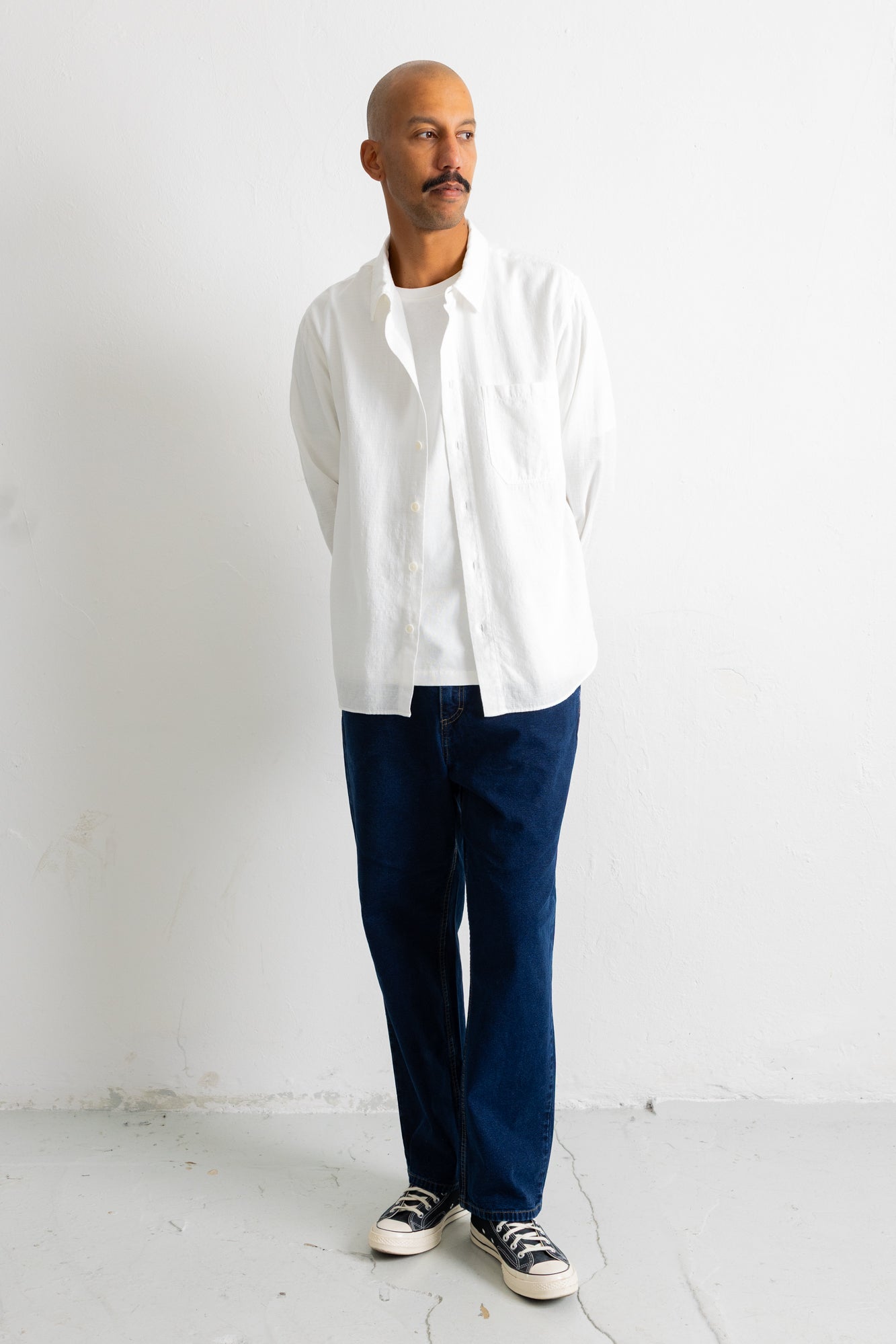 A man wearing a white long-sleeve button-up shirt, dark navy pants, and black-and-white sneakers, standing against a plain white wall.