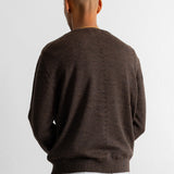 A man viewed from behind wearing a dark brown knit crewneck sweater and teal pants against a plain white wall.