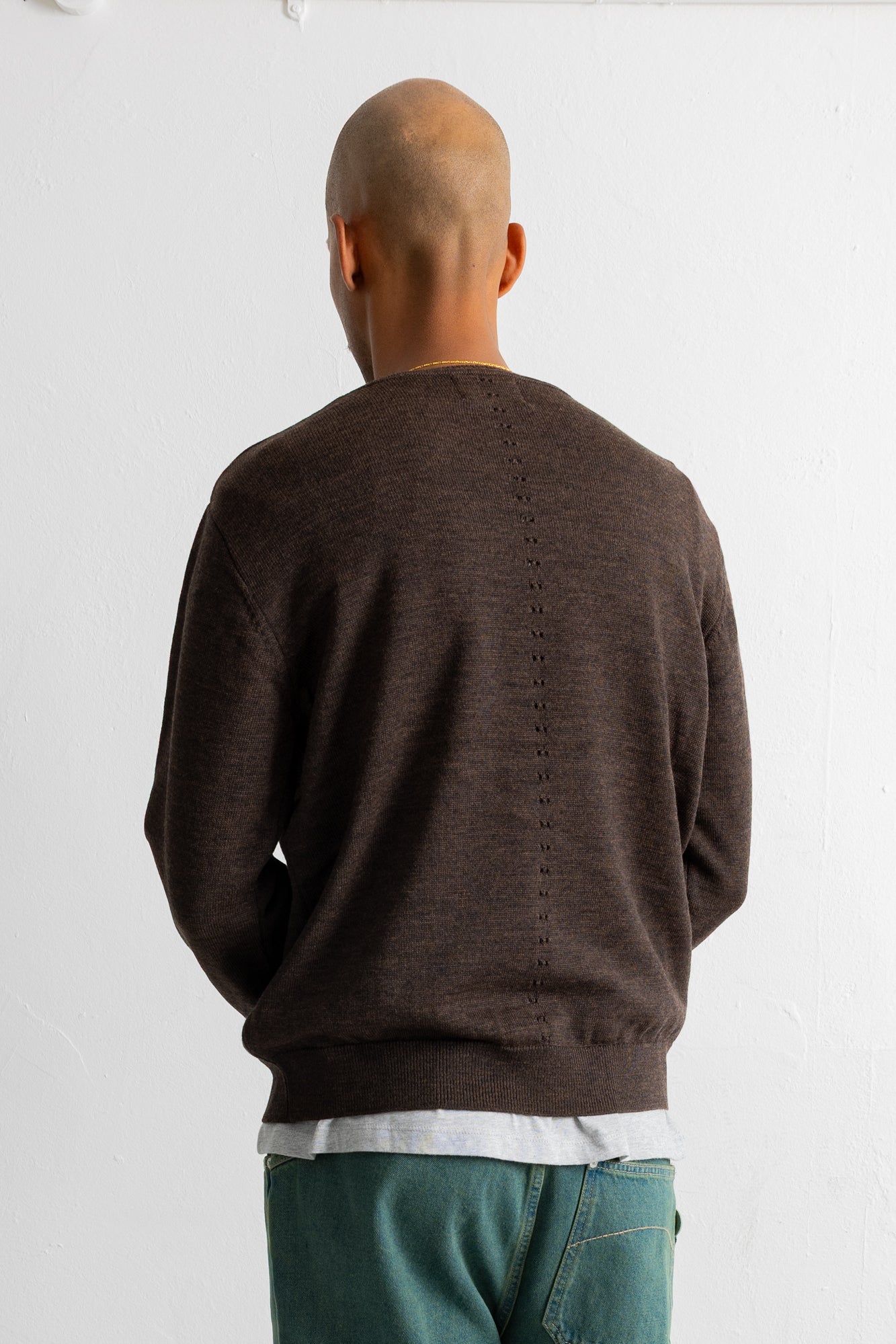 A man viewed from behind wearing a dark brown knit crewneck sweater and teal pants against a plain white wall.