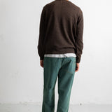 A man viewed from behind wearing a dark brown knit sweater, green trousers, and black-and-white sneakers, standing against a plain white wall.