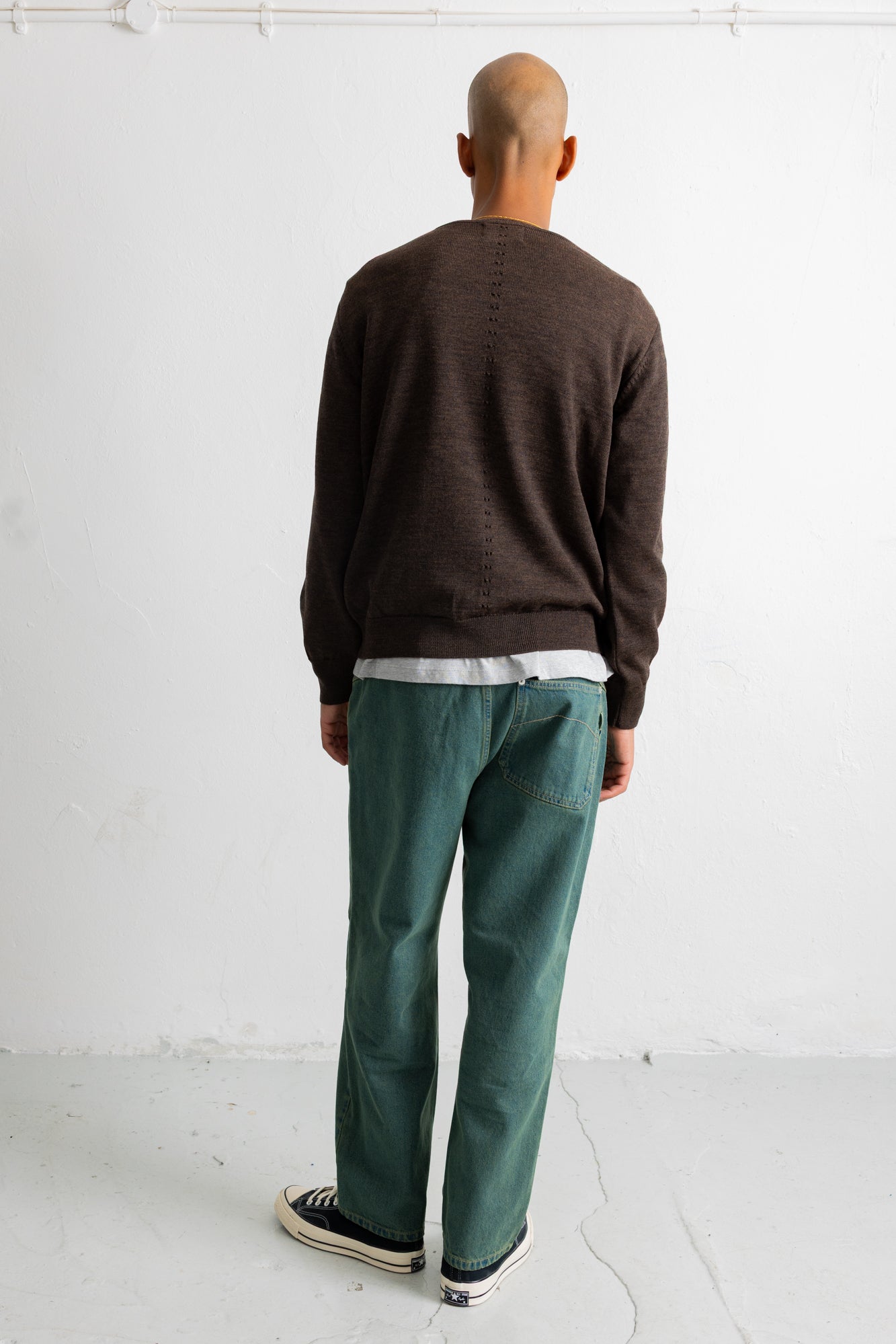 A man viewed from behind wearing a dark brown knit sweater, green trousers, and black-and-white sneakers, standing against a plain white wall.