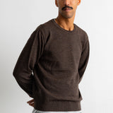 A man wearing a dark brown crewneck knit sweater and teal jeans, standing against a plain white wall.