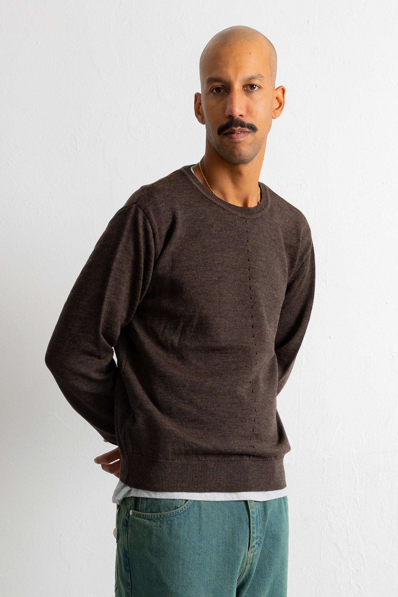 A man wearing a dark brown crewneck knit sweater and teal jeans, standing against a plain white wall.