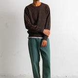 A man wearing a dark brown crewneck sweater, green trousers, and black canvas sneakers, standing against a white wall.