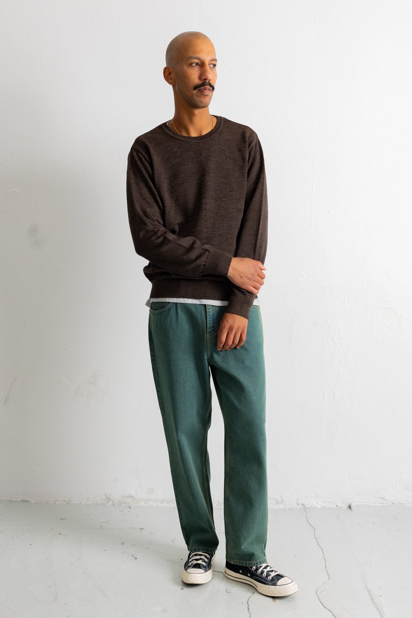 A man wearing a dark brown crewneck sweater, green trousers, and black canvas sneakers, standing against a white wall.