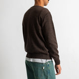 A man shown from the back wearing a dark brown knit sweater, a white undershirt, and teal jeans, standing against a plain light gray wall.