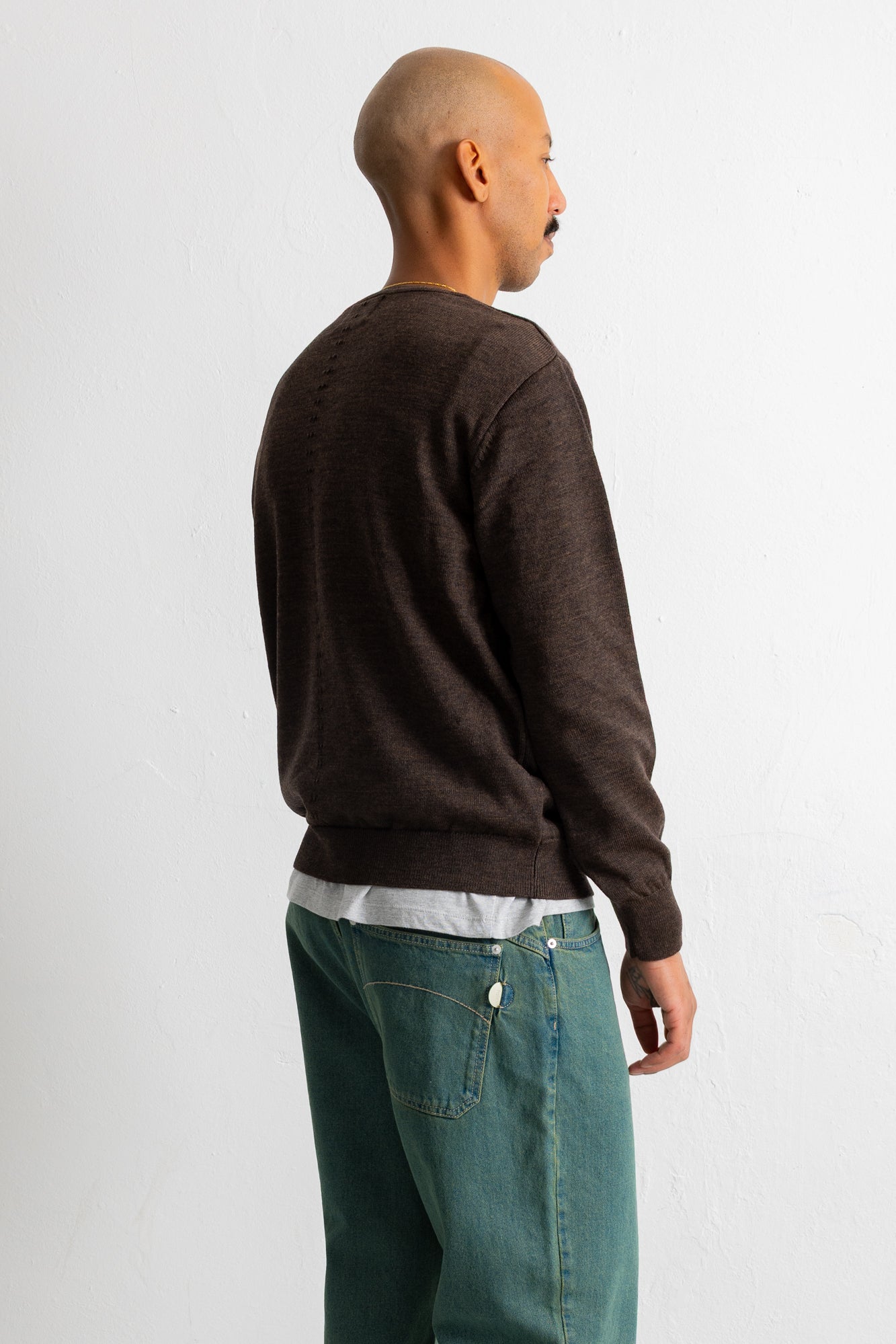 A man shown from the back wearing a dark brown knit sweater, a white undershirt, and teal jeans, standing against a plain light gray wall.