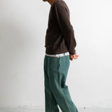 A man in profile wearing a dark brown long-sleeve sweater, teal wide-leg jeans, and black canvas sneakers, standing against a plain white wall.