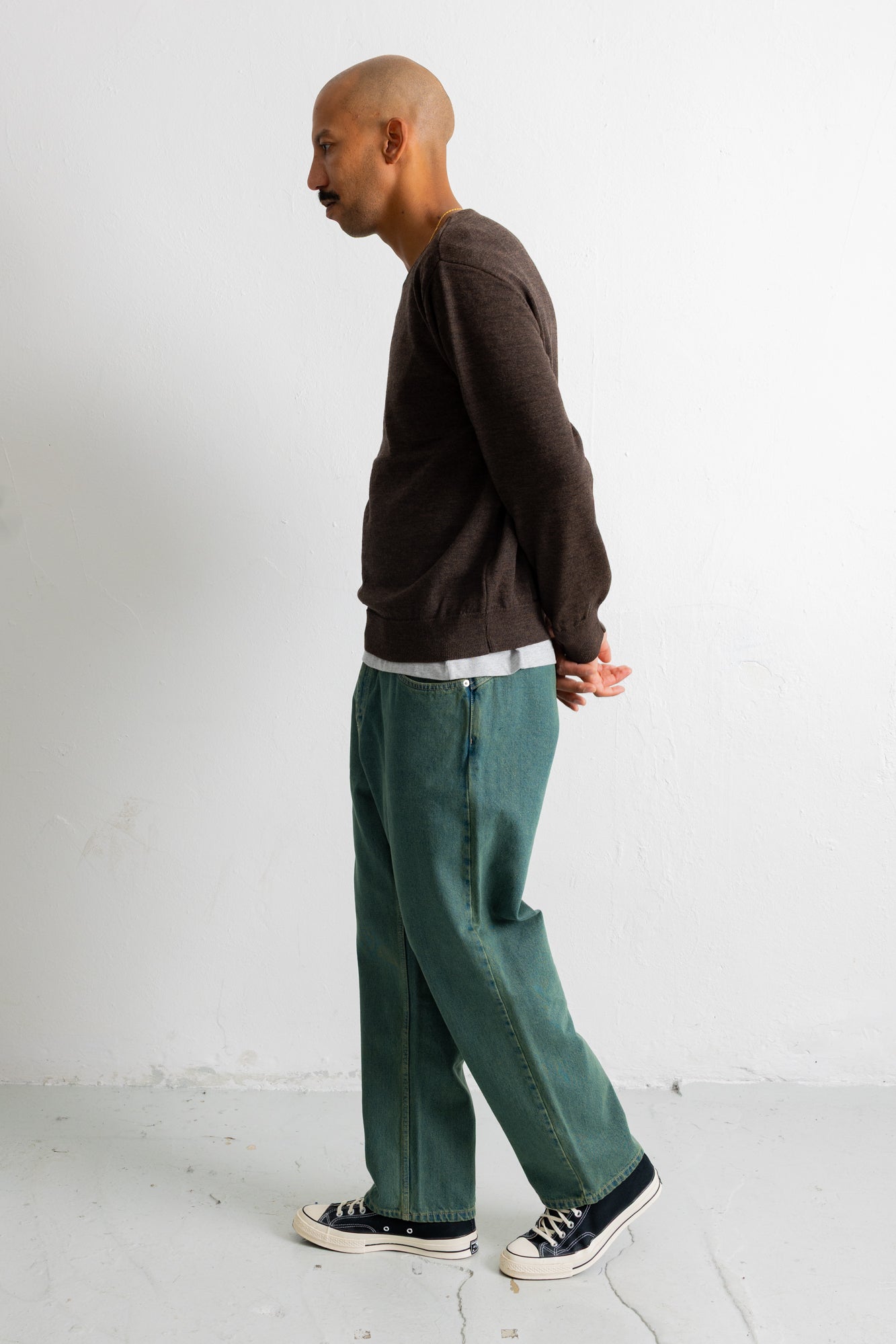 A man in profile wearing a dark brown long-sleeve sweater, teal wide-leg jeans, and black canvas sneakers, standing against a plain white wall.