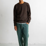 A man wearing a dark brown crewneck sweater, green trousers, and black and white sneakers, standing against a white studio wall.