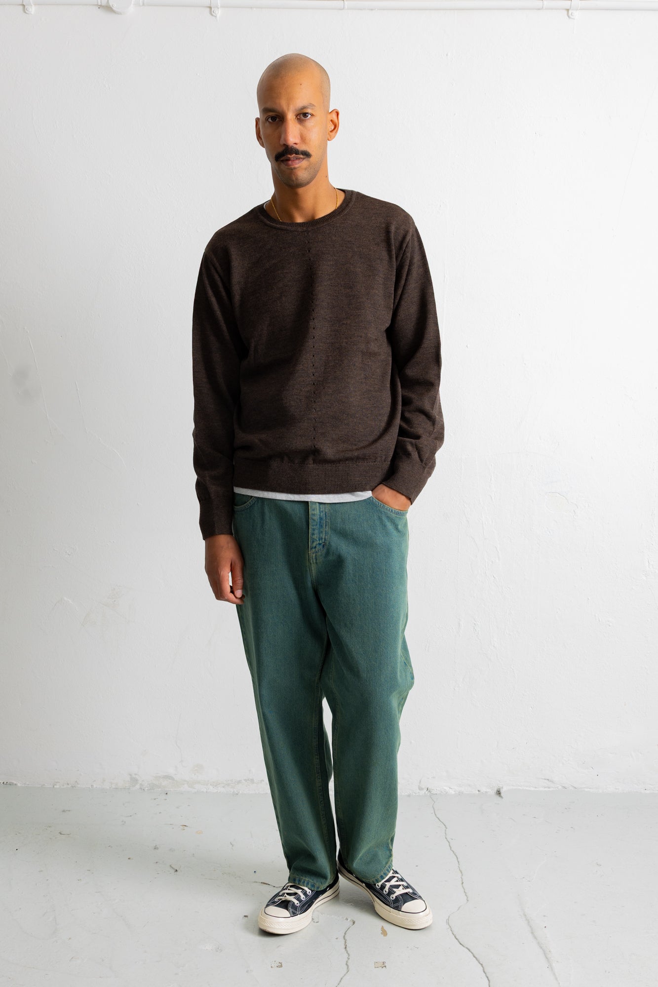 A man wearing a dark brown crewneck sweater, green trousers, and black and white sneakers, standing against a white studio wall.