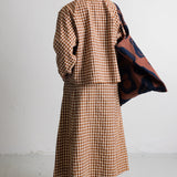 A model is shown from behind wearing a brown and beige gingham long-sleeve top and matching midi skirt, with a dark blue and rust patterned cardigan draped over one shoulder, set against a plain white wall.