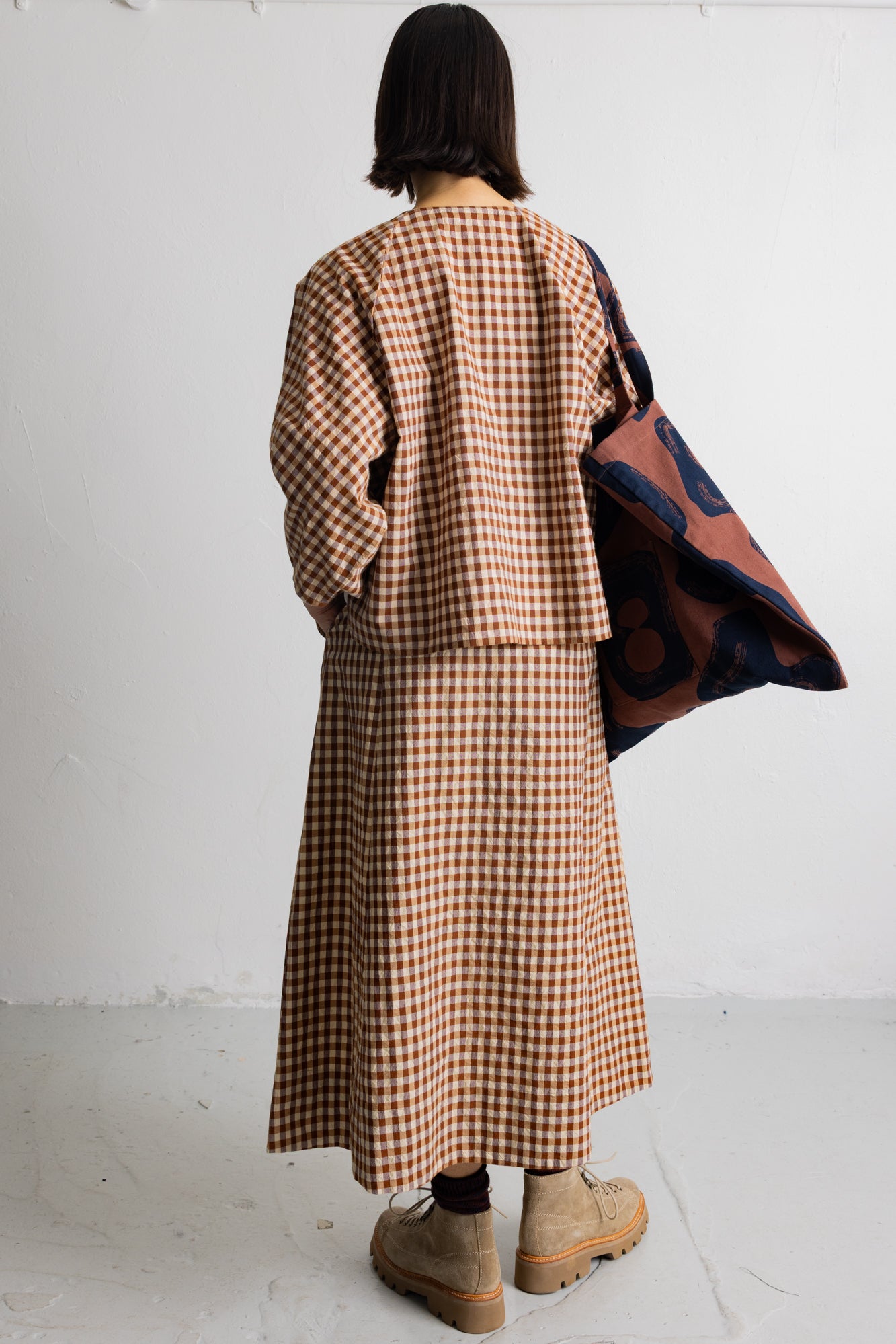 A model is shown from behind wearing a brown and beige gingham long-sleeve top and matching midi skirt, with a dark blue and rust patterned cardigan draped over one shoulder, set against a plain white wall.