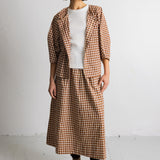 A model stands against a white studio wall wearing a brown and beige gingham check blazer and matching midi skirt over a white top, paired with beige lace-up boots.