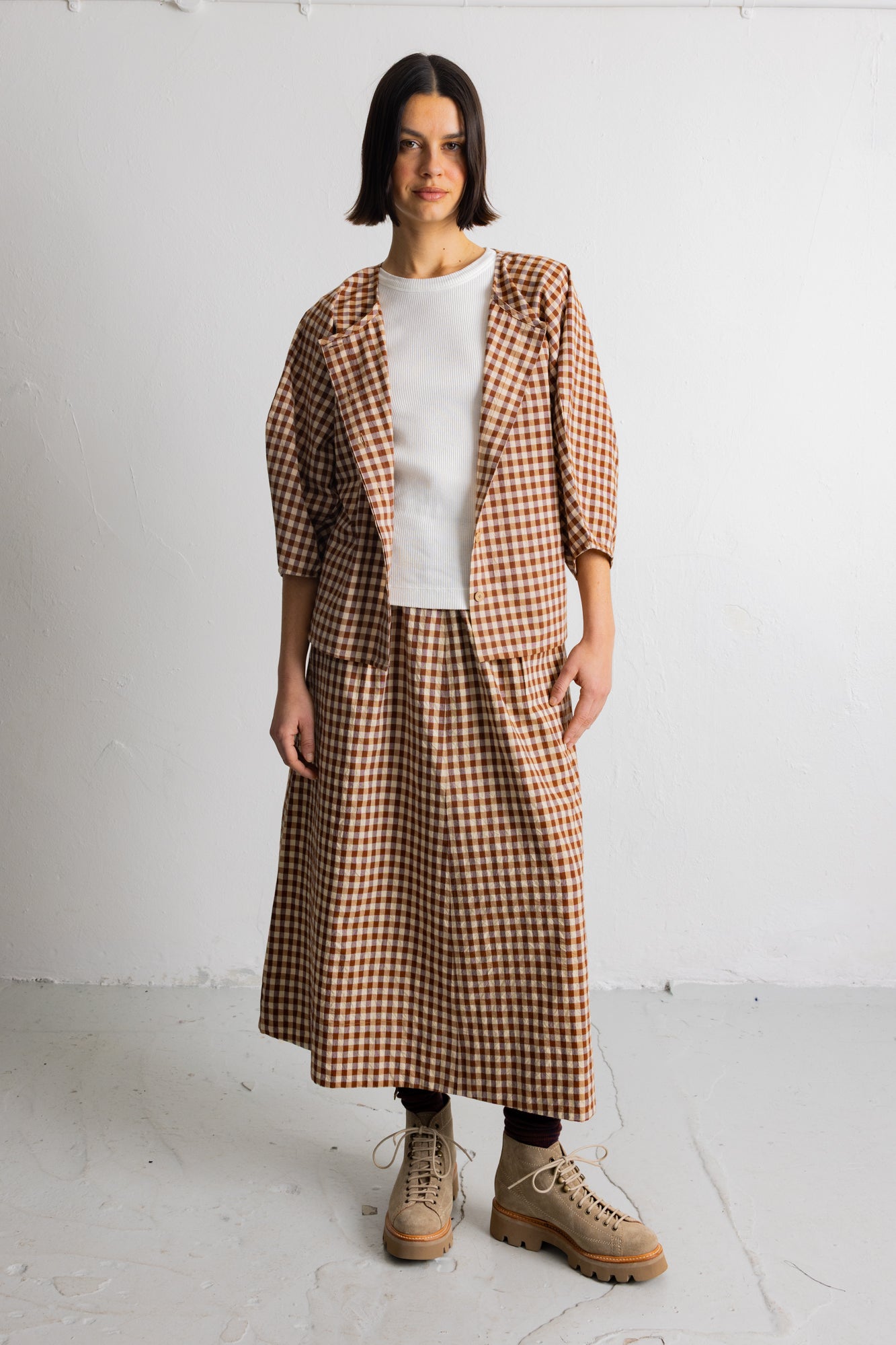A model stands against a white studio wall wearing a brown and beige gingham check blazer and matching midi skirt over a white top, paired with beige lace-up boots.
