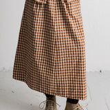 A person wearing a brown and beige gingham check midi skirt with a matching blazer, a white top, and beige lace-up boots, standing against a white wall.