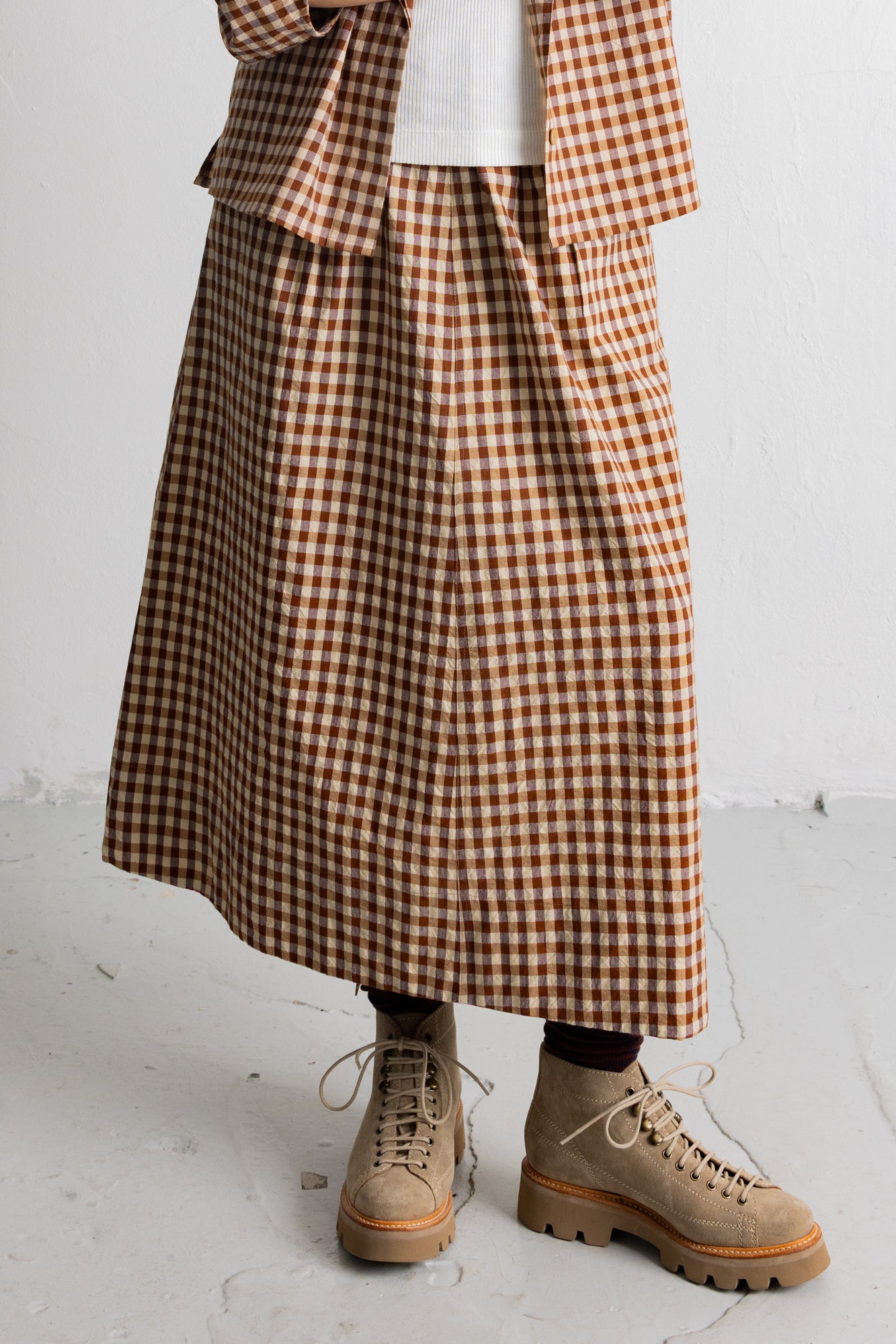 A person wearing a brown and beige gingham check midi skirt with a matching blazer, a white top, and beige lace-up boots, standing against a white wall.