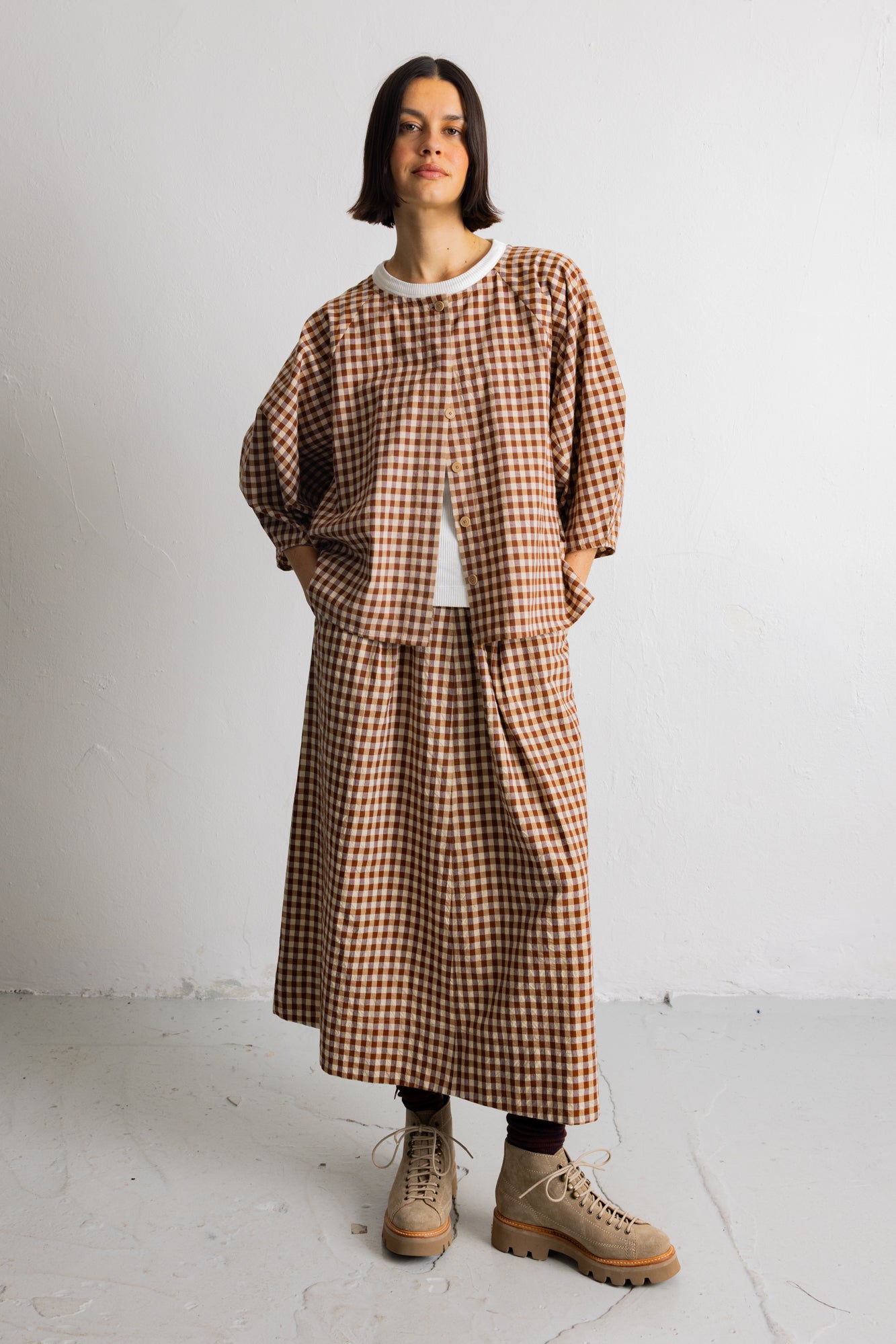 A model stands against a plain white wall wearing a brown and beige gingham check long-sleeve top and matching skirt set, styled with a white undershirt, dark socks, and beige lace-up boots.