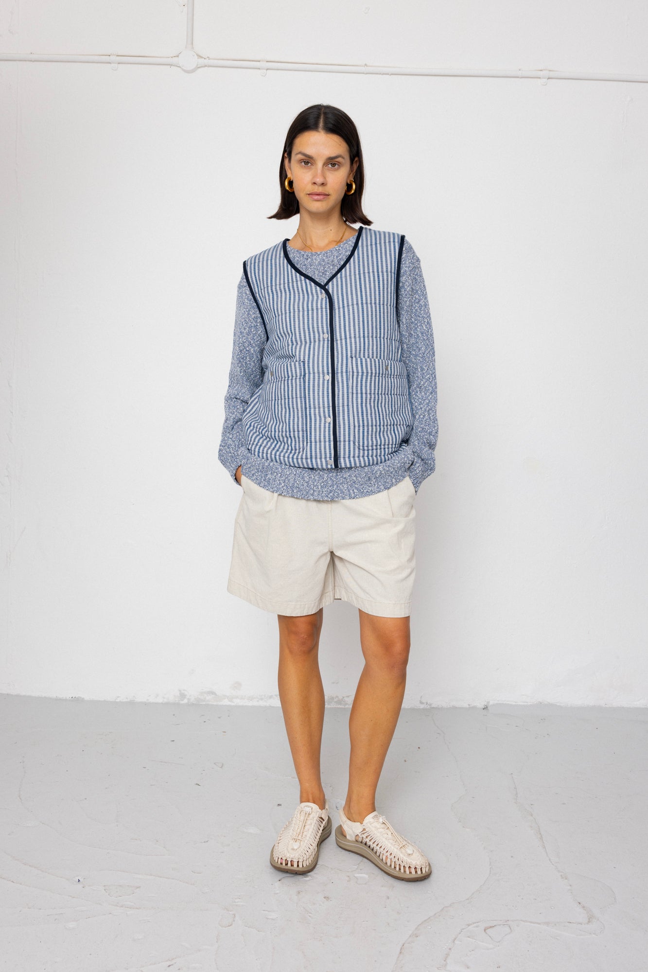 Person wearing a blue striped vest over a blue jumper and white shorts against a white background 